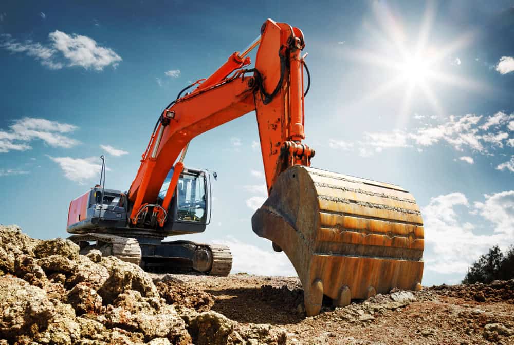 A large orange excavator with a metal bucket is digging into rocky soil under a bright blue sky with scattered clouds. The sun shines brightly from the top right corner of the image, casting shadows on the construction site where Septic Tank Services Suffolk County are being performed.