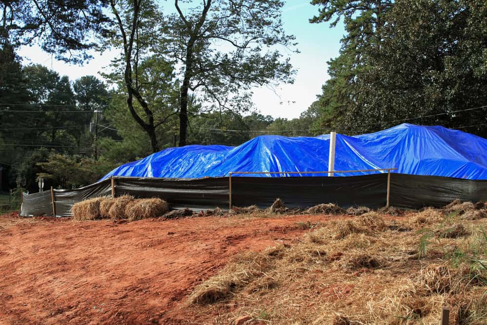 A construction site with a ground covered in red dirt and surrounding straw features a structure being built under large blue tarps, with a black barrier along the perimeter. Tall trees stand in the background, hinting at upcoming cesspool maintenance services in Suffolk County.