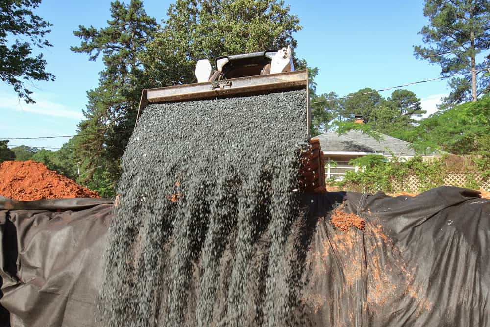 A construction scene where gravel is being poured from a large metal bucket onto a black tarp. Trees and a house are visible in the background, hinting at the need for Cesspool Services Suffolk County amidst ongoing improvements.