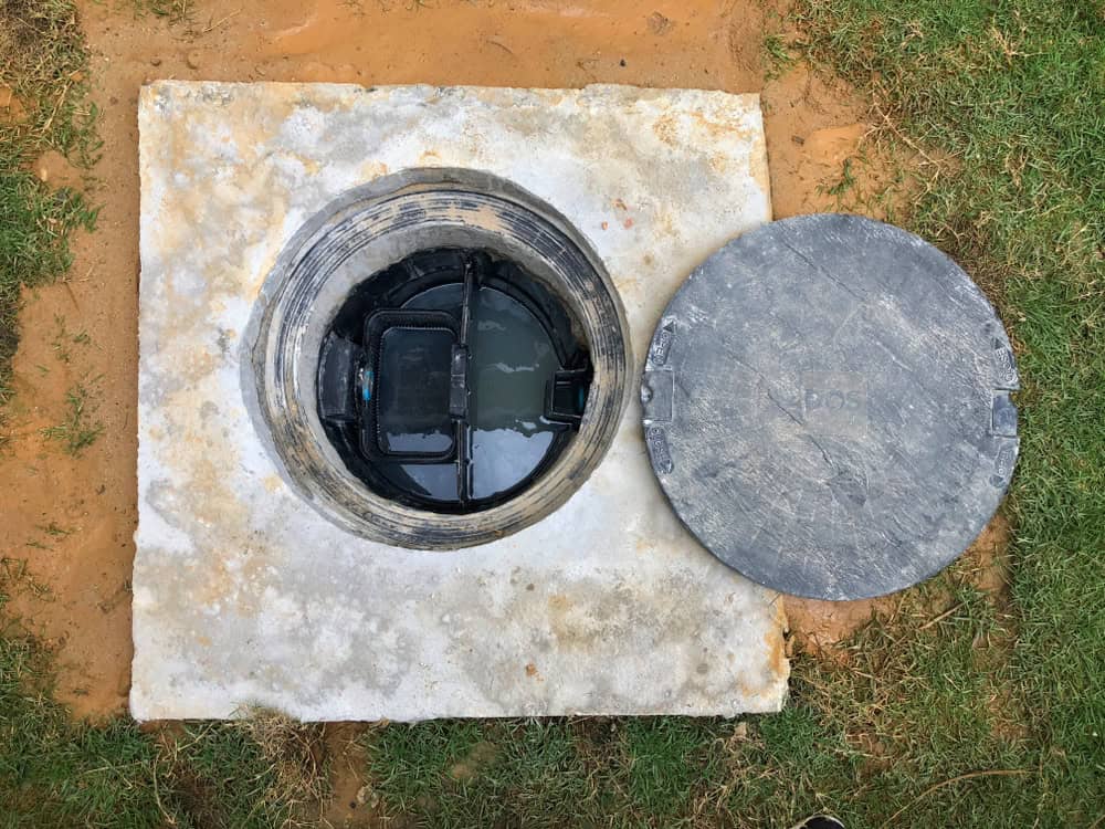 A concrete septic tank with its cover removed, revealing the water and internal components inside. The round, detached cover lies on the grass beside the tank opening. Surrounding the tank is a patch of uneven grass and exposed soil, highlighting signs of recent septic tank pumping.