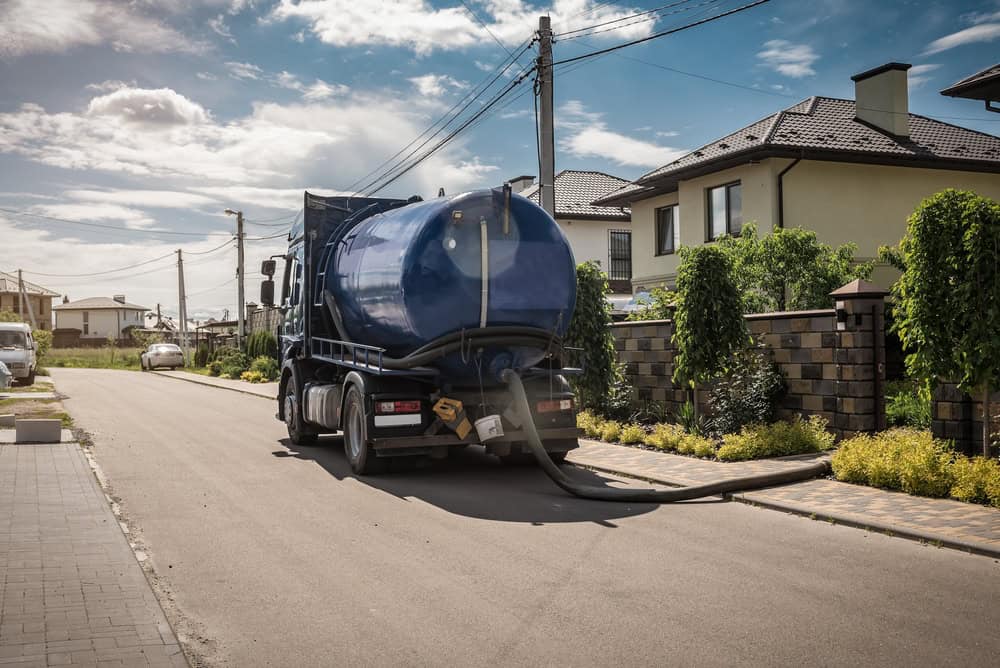 A blue septic tank truck is parked on the side of a residential street in a suburban neighborhood on a sunny day, ready for its next job in cesspool cleaning. A hose extends from the back of the truck onto the sidewalk. Modern houses with well-maintained gardens are visible in the background.