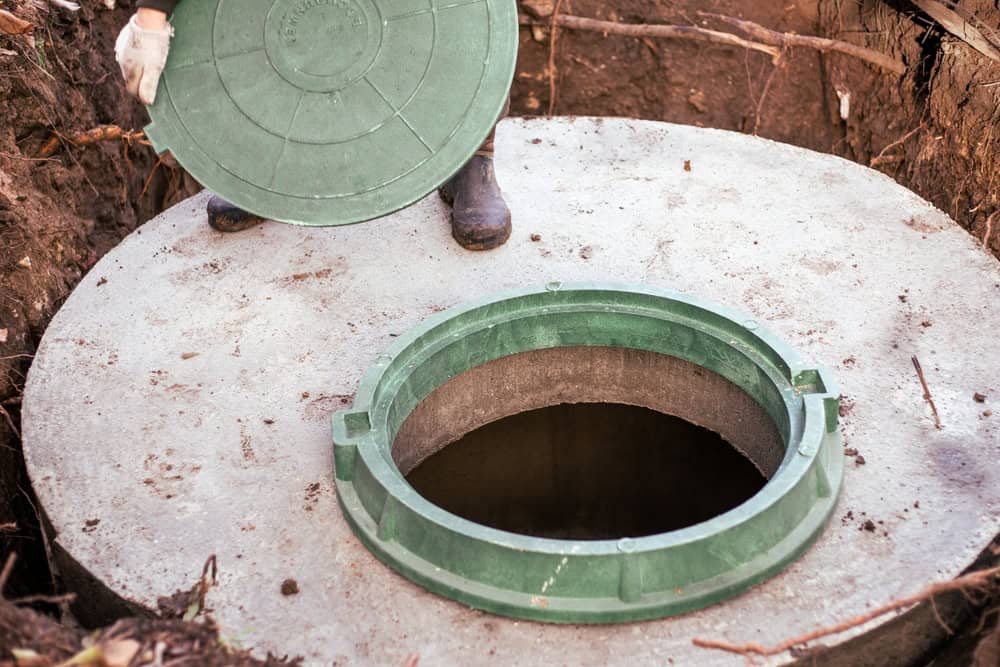 A person wearing gloves and boots lifts the green lid off an underground concrete septic tank. The circular opening of the septic tank, ideal for cesspool cleaning, is surrounded by dirt and roots.