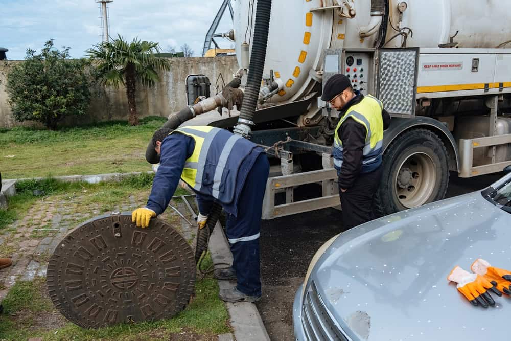 Two utility workers, dressed in high-visibility vests, gloves, and beanies, are removing a manhole cover. A sewage vacuum truck is parked nearby with hoses connected to the cesspool. A gray car is partially visible in the foreground, set against an urban environment with trees and a fence.