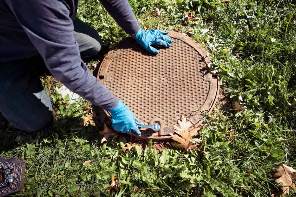 A person wearing blue gloves is kneeling on grass and using a wrench to open a round metal manhole cover, preparing for cesspool cleaning. Leaves and greenery are scattered around on the ground, while a set of sockets lies near the edge of the frame.