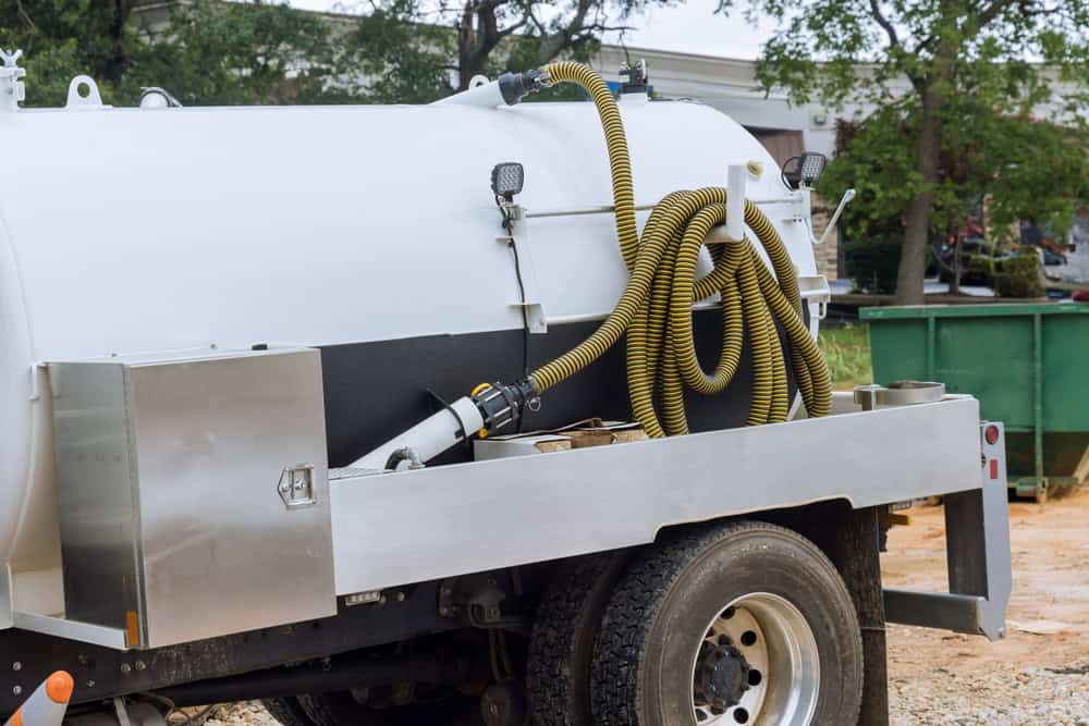 A close-up view of a large, white industrial vacuum truck parked on a dirt surface near a green dumpster, with a yellow hose attached to the side. This powerful vehicle is perfect for cesspool cleaning. The background features trees and a building.